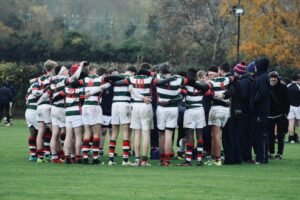 Rugby players in striped jerseys huddle during a game on an autumn field.